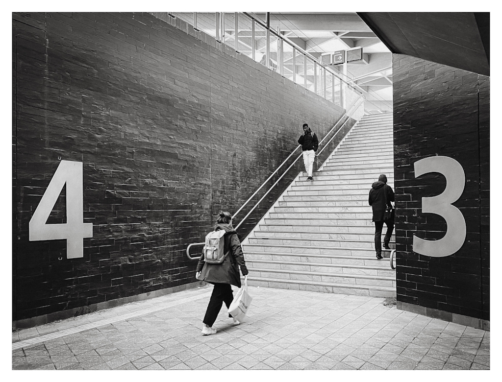 People walking up or down the stairs at a train station, with the number 4 and 3 indicating the platforms shown on the wall. 