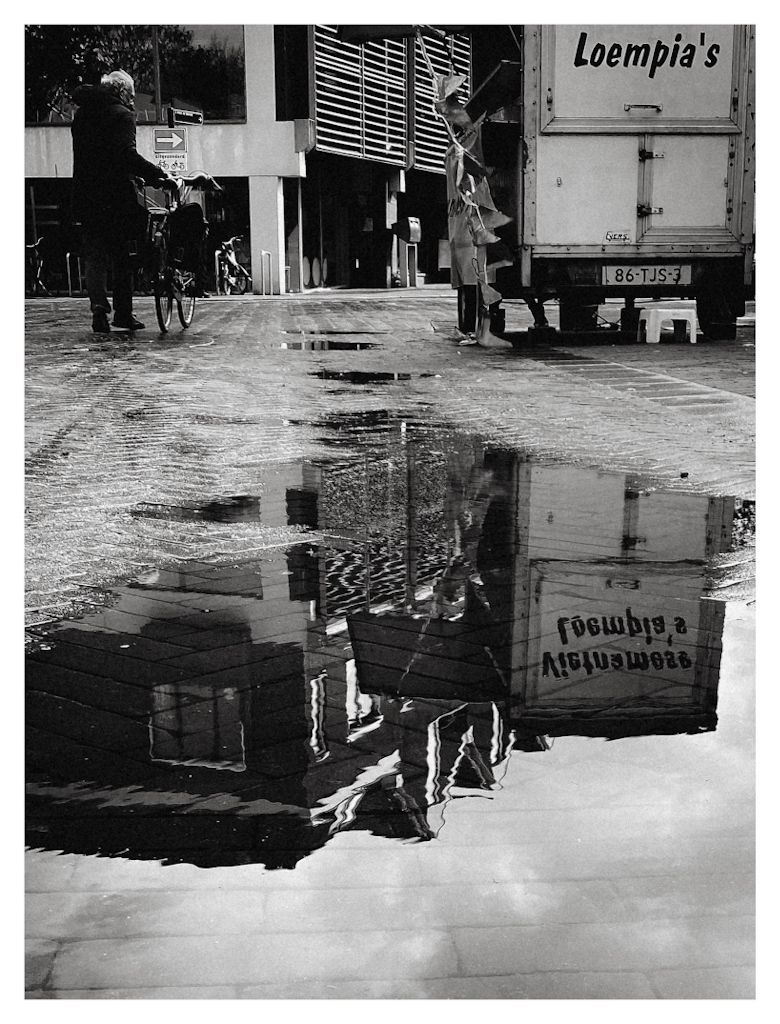 Street stall with the text ‘Vietnamese loempia’s’ on it reflected in a puddle. And a man with a bicycle walking on the street. &10;