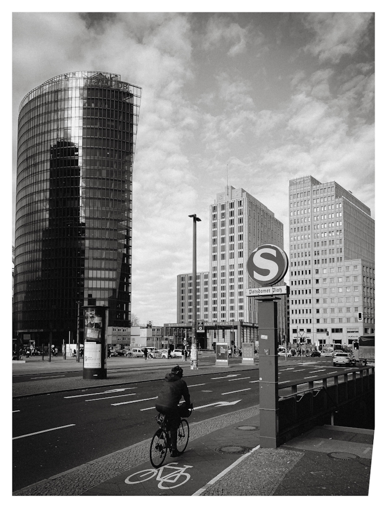 Cyclist next to the S-bahn sign at Potsdamer Platz, with the empty road and high buildings in the background. 