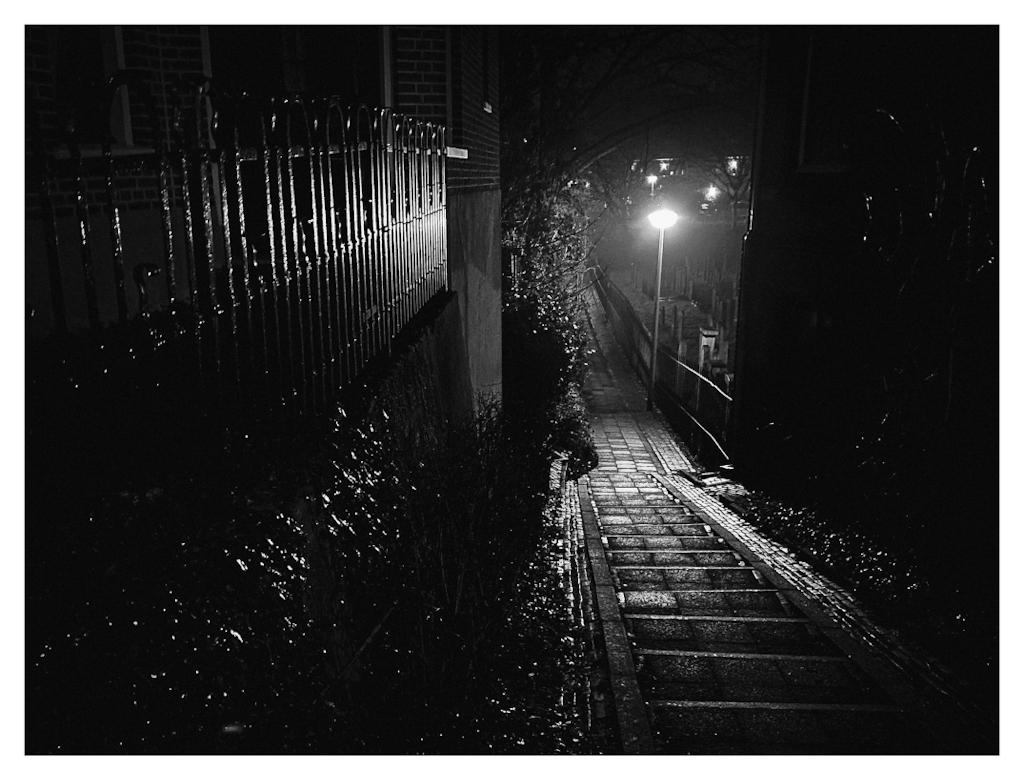 Diagonal lines of a fence and stairs in a dimly lit street scene, in B&W. 